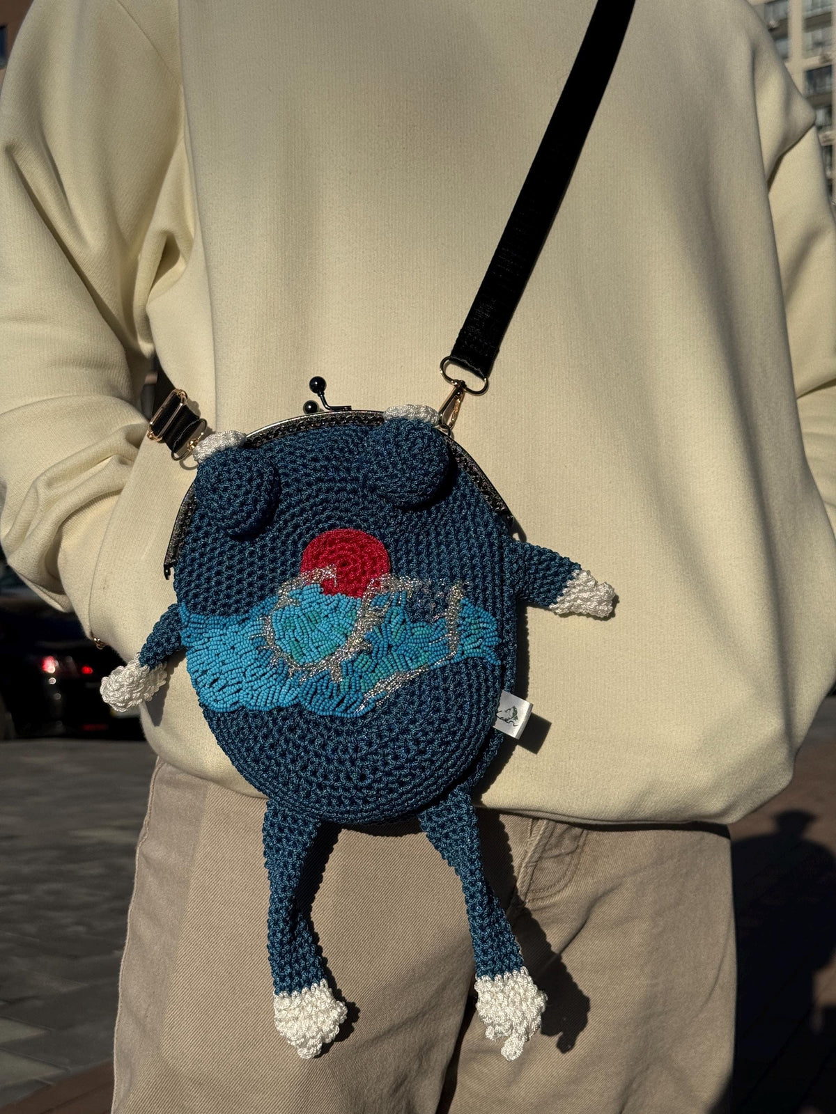 Dark blue crochet frog bag with wave and red sun embroidery, worn crossbody on a person in a cream sweater