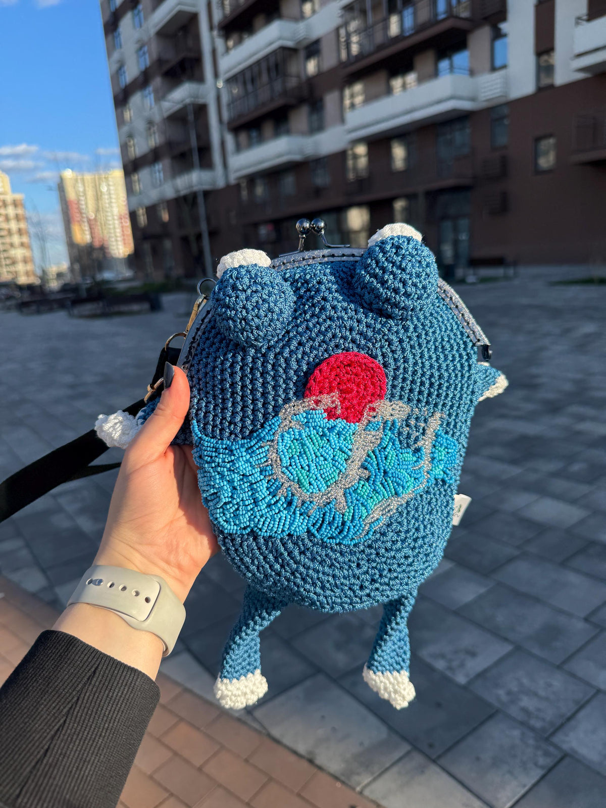 Blue crochet frog bag with embroidered wave and red sun motif, inspired by Japanese aesthetics, held in hand against a backdrop of modern urban buildings