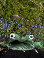 Hand holding a crochet frog bag in greenery with visible mouth