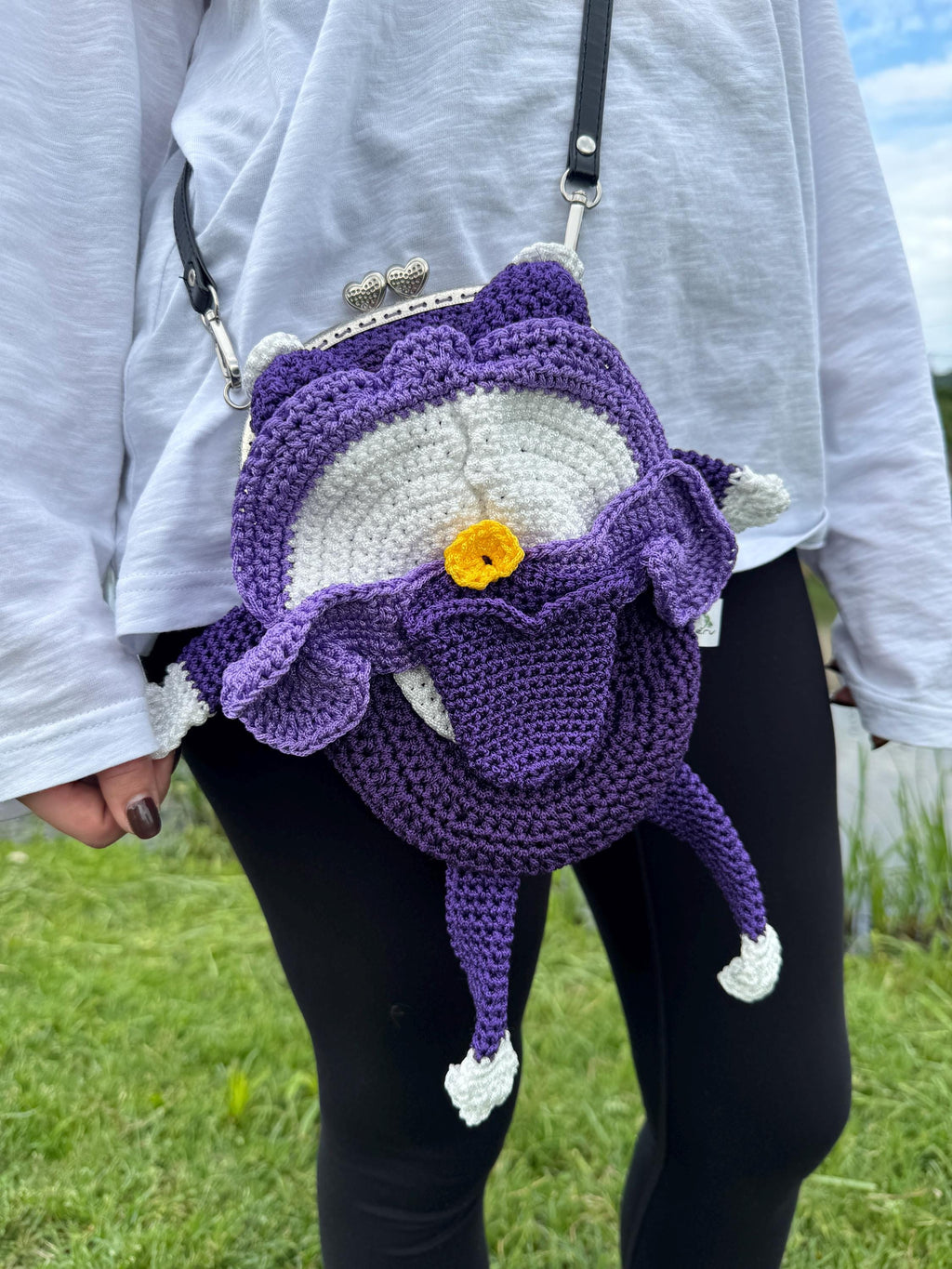Purple crochet frog bag with frilly details and a yellow flower on the belly, worn crossbody on a person in a white top and black pants
