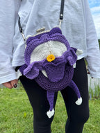 Purple crochet frog bag with frilly details and a yellow flower on the belly, worn crossbody on a person in a white top and black pants