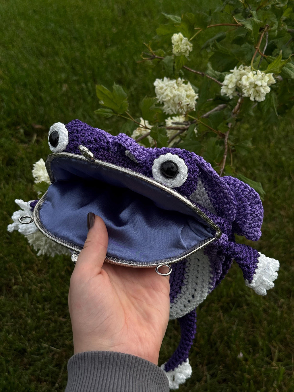 Hand holding an open purple crochet frog bag with light blue-gray lining and clasp frame, with flowers and greenery in the background