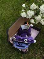 Purple crochet frog bag with open mouth and blue lining, presented in a gift box with tag and tissue paper, placed on grass near blooming white flowers