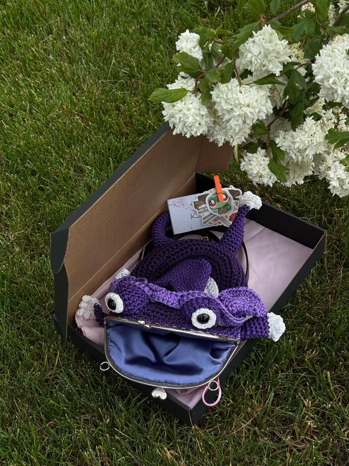 Purple crochet frog bag with open mouth and blue lining, presented in a gift box with tag and tissue paper, placed on grass near blooming white flowers