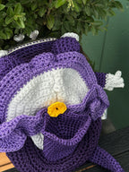 Back view of purple crochet frog bag featuring an embroidered violet flower with a yellow center on its back, displayed in front of green leaves
