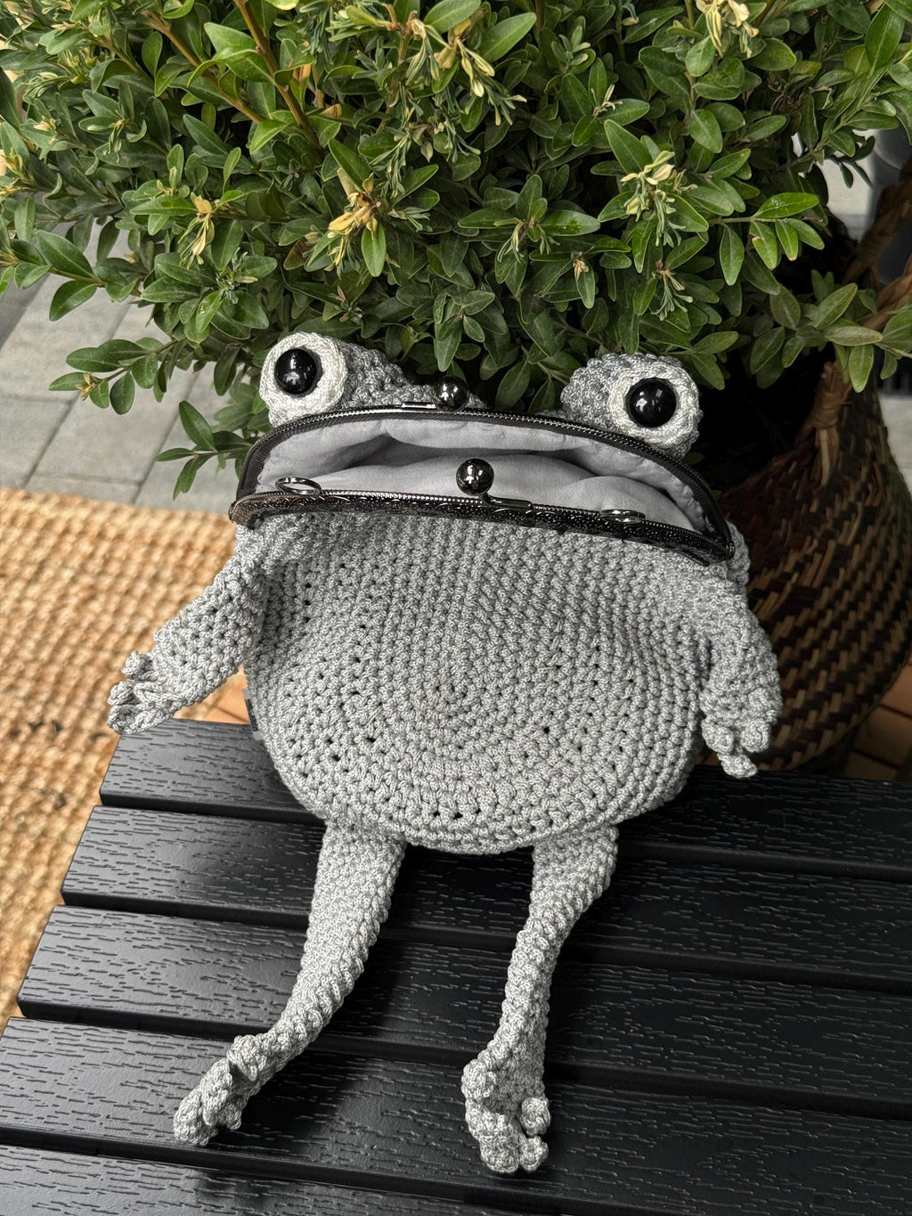 Gray crochet frog bag with bulging eyes and long limbs, sitting on a dark bench in front of green leaves