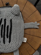 Side view of gray crochet frog bag with black embroidery and outstretched arm, displayed on a wooden surface