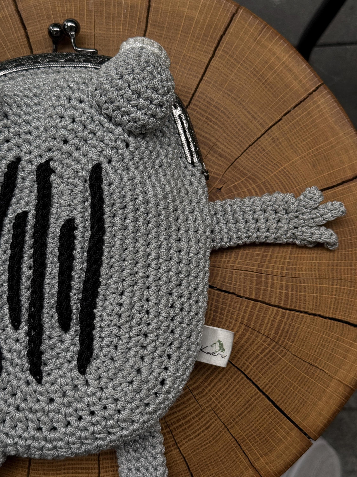 Side view of gray crochet frog bag with black embroidery and outstretched arm, displayed on a wooden surface