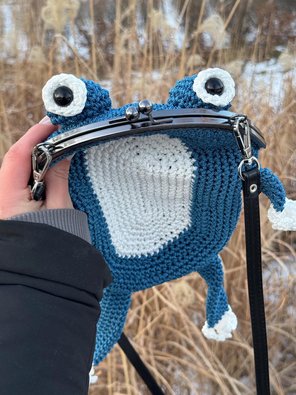 Front view of blue crochet frog bag with white belly and metal clasp, held in hand against a backdrop of dry winter grass