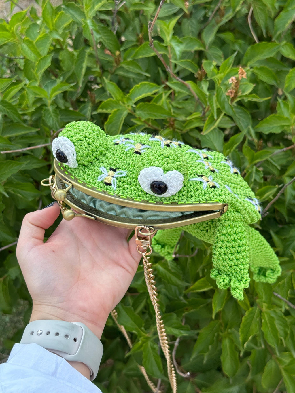 Side view of a green crochet frog purse with big round eyes and a gold kiss clasp. The frog bag features embroidered bees and a detachable gold chain strap. Whimsical handmade animal bag shaped like a frog.