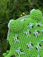 Top view of a handmade crochet frog bag in bright green color, decorated with embroidered bees. The frog purse has four legs and rounded eyes, displayed against green leaves. Unique animal-shaped bag for frog and bee lovers.
