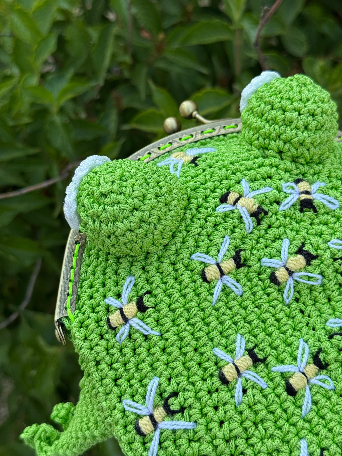 Top view of a handmade crochet frog bag in bright green color, decorated with embroidered bees. The frog purse has four legs and rounded eyes, displayed against green leaves. Unique animal-shaped bag for frog and bee lovers.